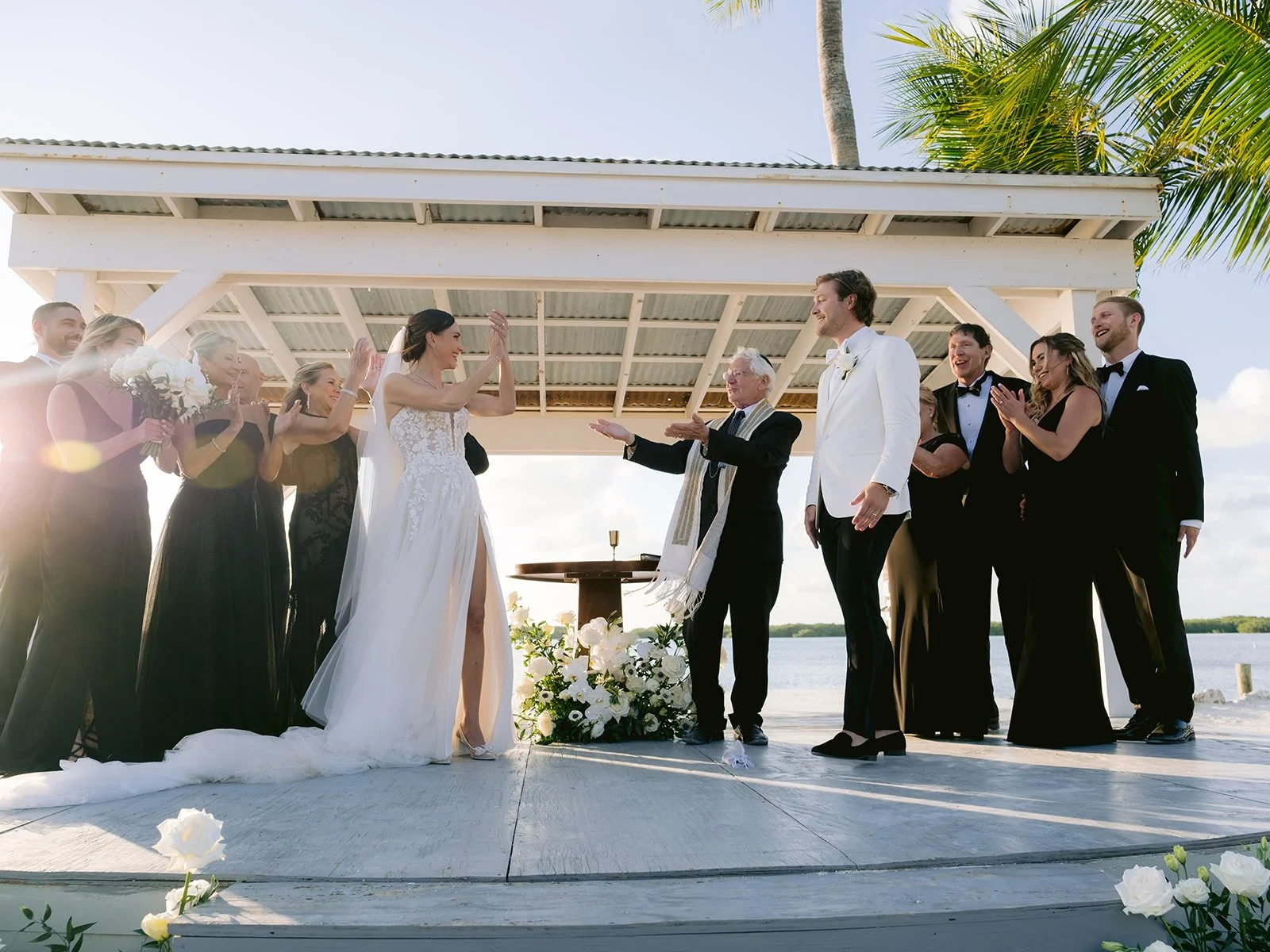 A wedding ceremony outdoors with a bride in a white dress and a groom in a white jacket, surrounded by a bridal party in formal attire, under a gazebo by the water.