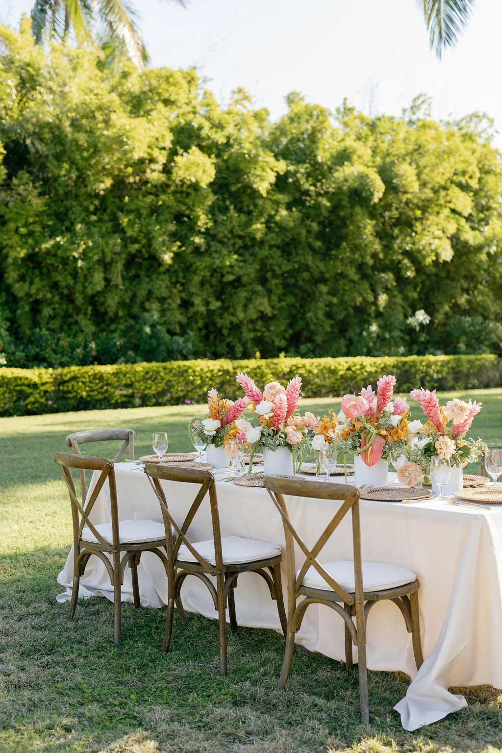 A long banquet table with a white tablecloth and floral centerpieces, set outdoors with chairs around it, surrounded by greenery and trees.