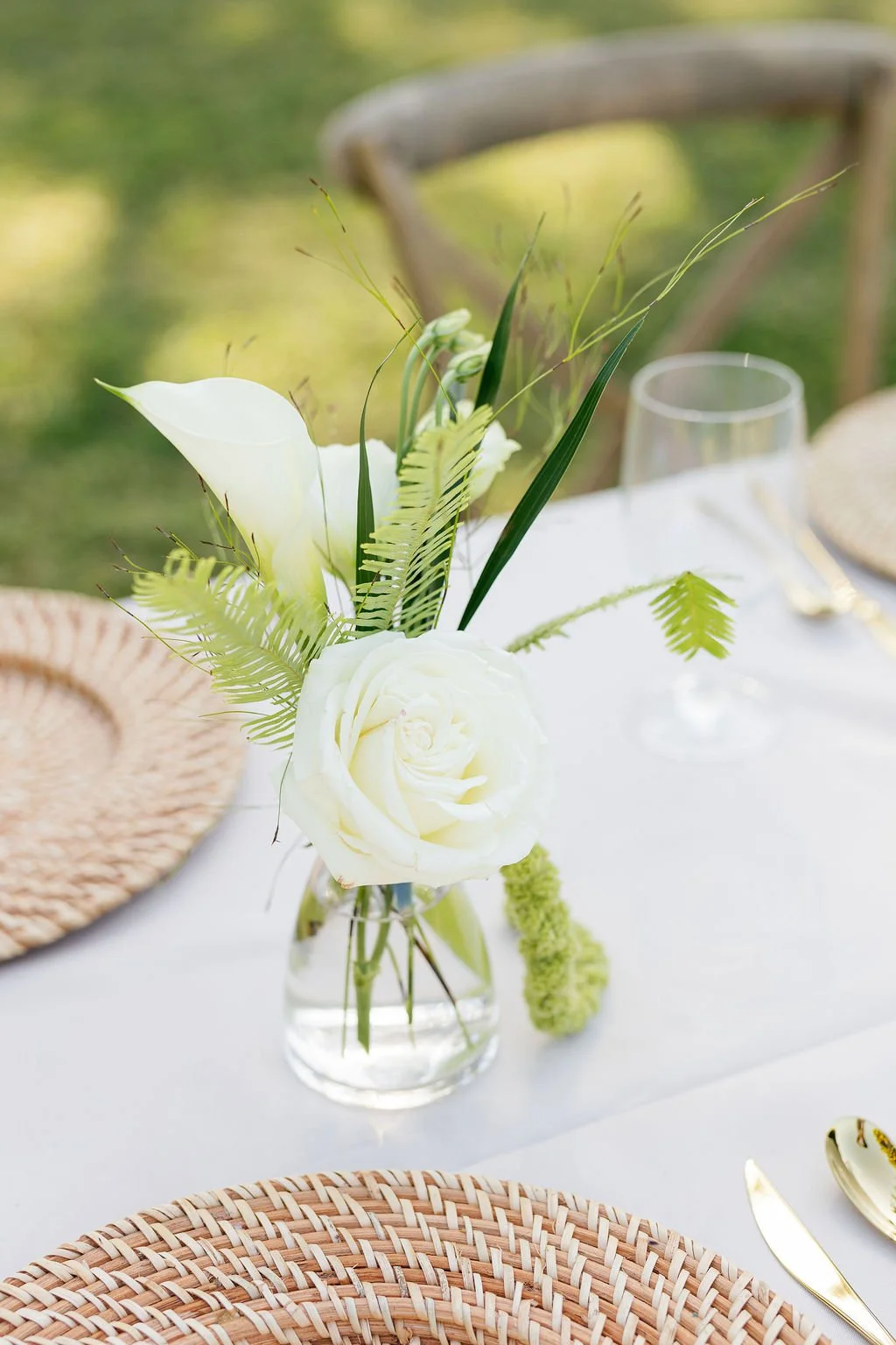 White floral centerpiece with roses, calla lilies, ferns, and greenery on a white tablecloth, set for an outdoor event with glassware and woven placemats.