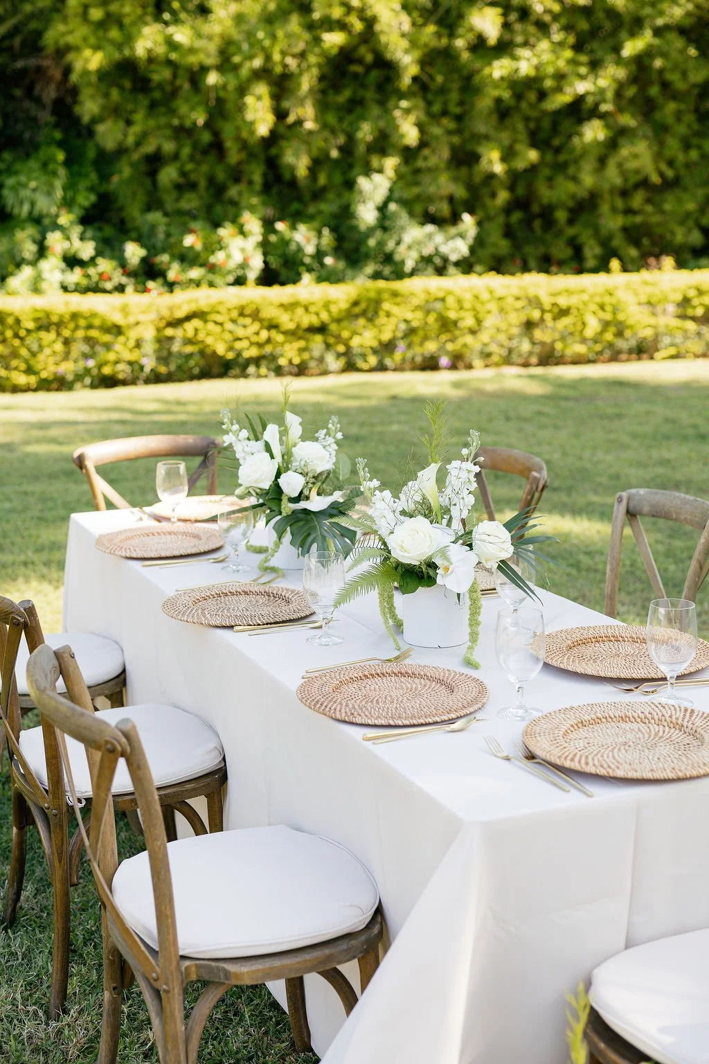 Outdoor banquet table decorated with white floral centerpieces, wicker placemats, glassware, and gold flatware, set on a grassy area with lush green trees and bushes in the background.