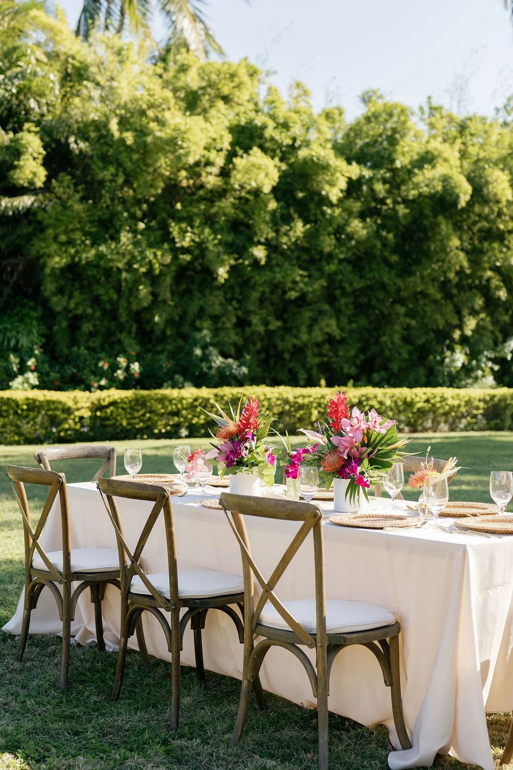 An outdoor dining table set with tropical pink and red flower arrangements in white vases, glassware, and woven placemats, situated on a grassy area with trees and a hedge in the background.