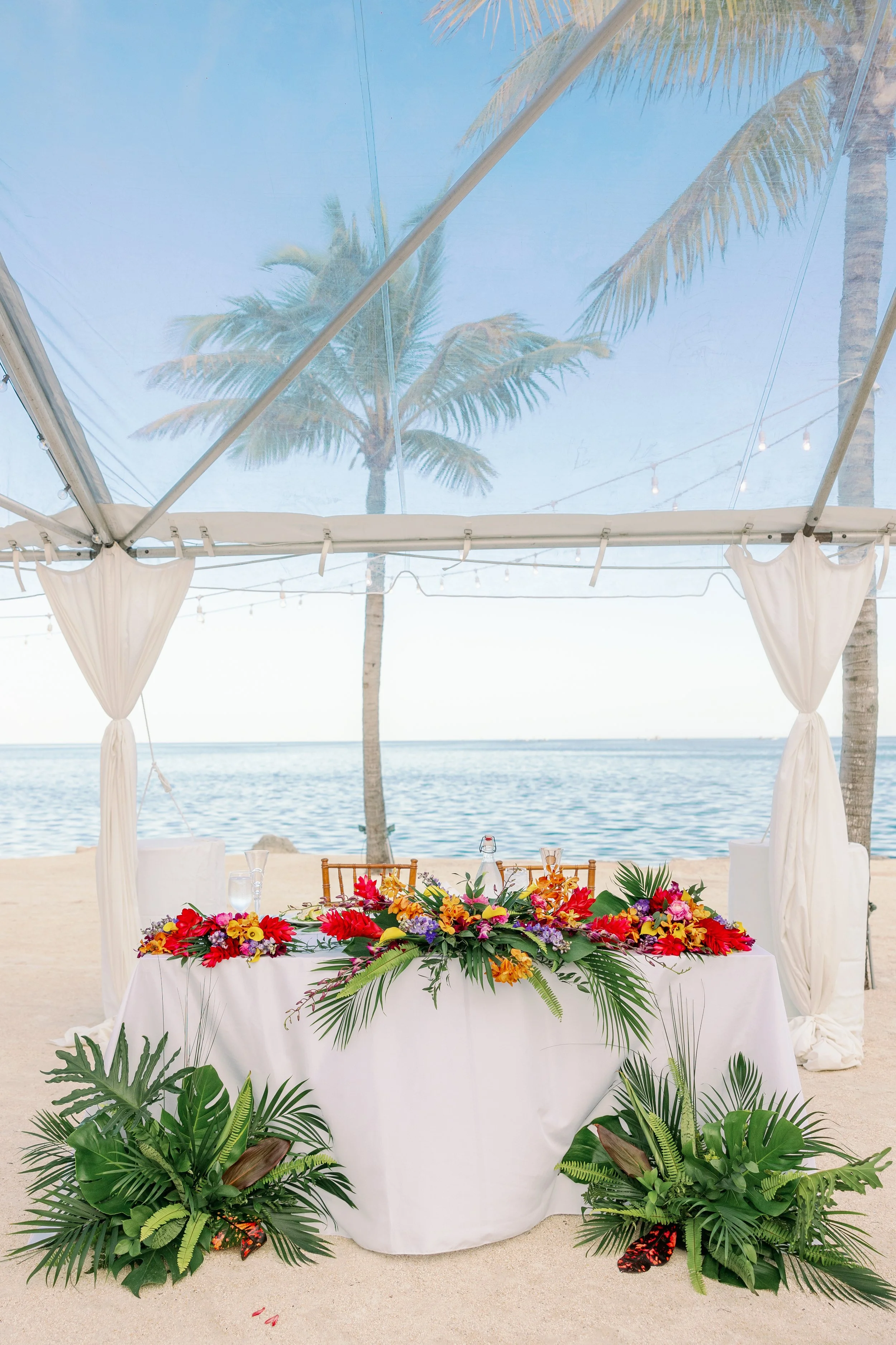 Beachfront wedding or event setup under a clear tent, with a white-draped table adorned with vibrant tropical flowers and greenery. Palm trees and ocean visible in the background.