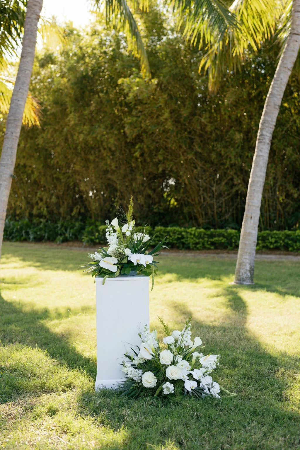 A white floral arrangement with white roses, orchids, and greenery on a white pedestal in an outdoor garden with palm trees and lush green bushes.