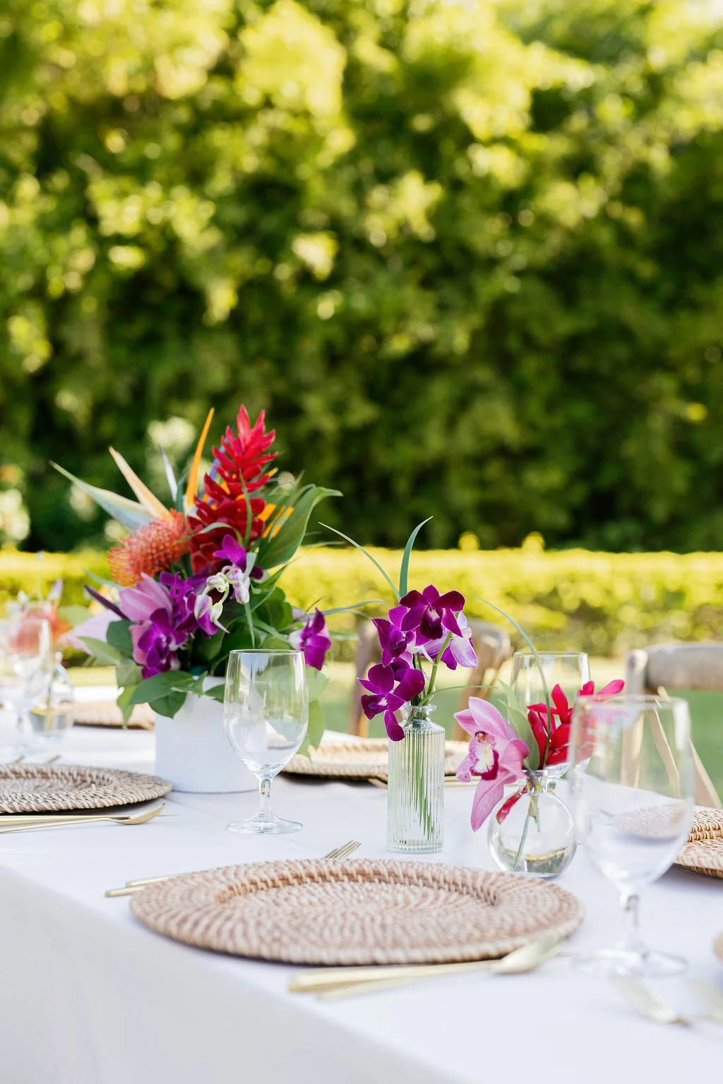 Elegant outdoor table setting with floral centerpieces, wine glasses, woven placemats, and gold utensils, set against a lush green garden backdrop.