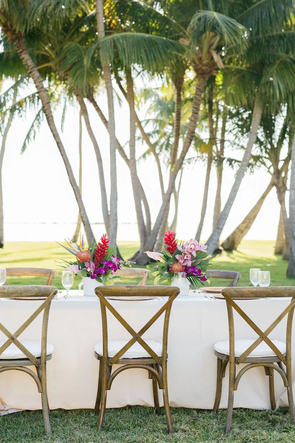 Outdoor dining table set with floral arrangements and wine glasses, with palm trees in the background.