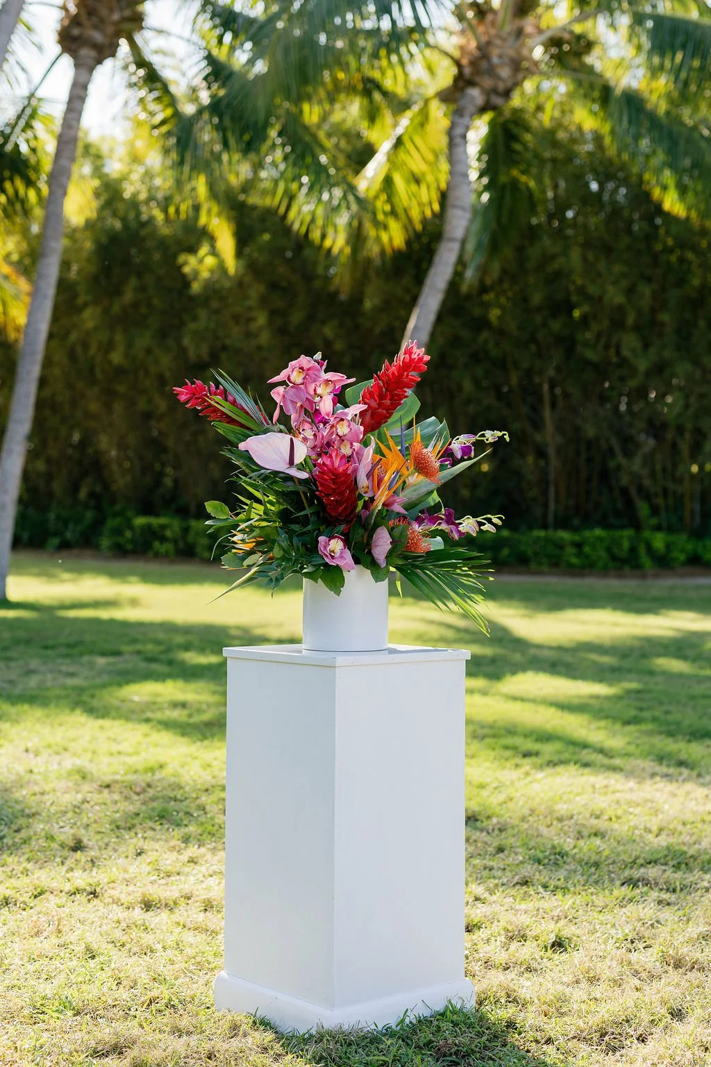 Colorful tropical flower arrangement in a white vase on a white pedestal outdoors, with palm trees and green foliage in the background.