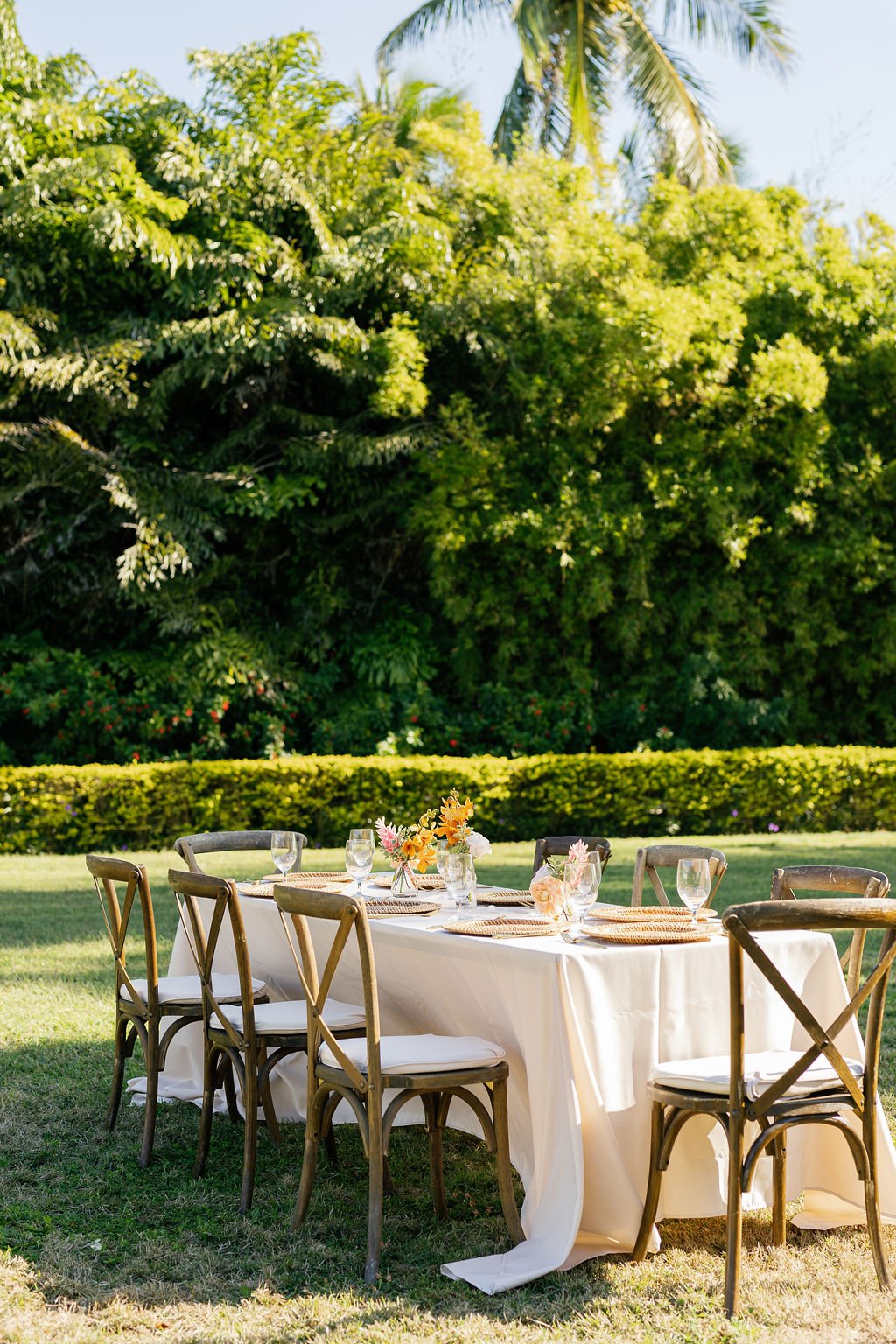 An outdoor dining table set for a gathering with six wooden chairs, decorated with a white tablecloth, floral centerpieces, and glassware, in a lush green garden with trees and bushes in the background.