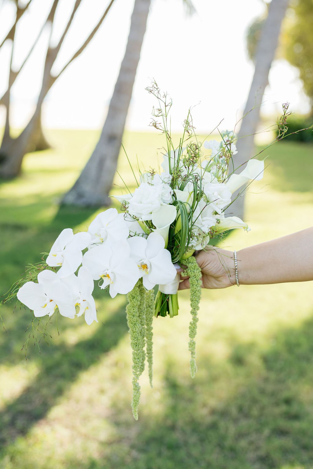 A person's hand holding a bouquet of white flowers, including orchids and greenery, outdoors with trees and sunlight in the background.
