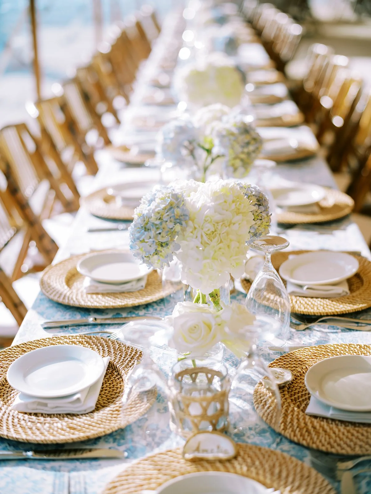 Elegant outdoor dining table setting with woven placemats, white plates, glassware, and floral centerpieces featuring white and blue hydrangeas.