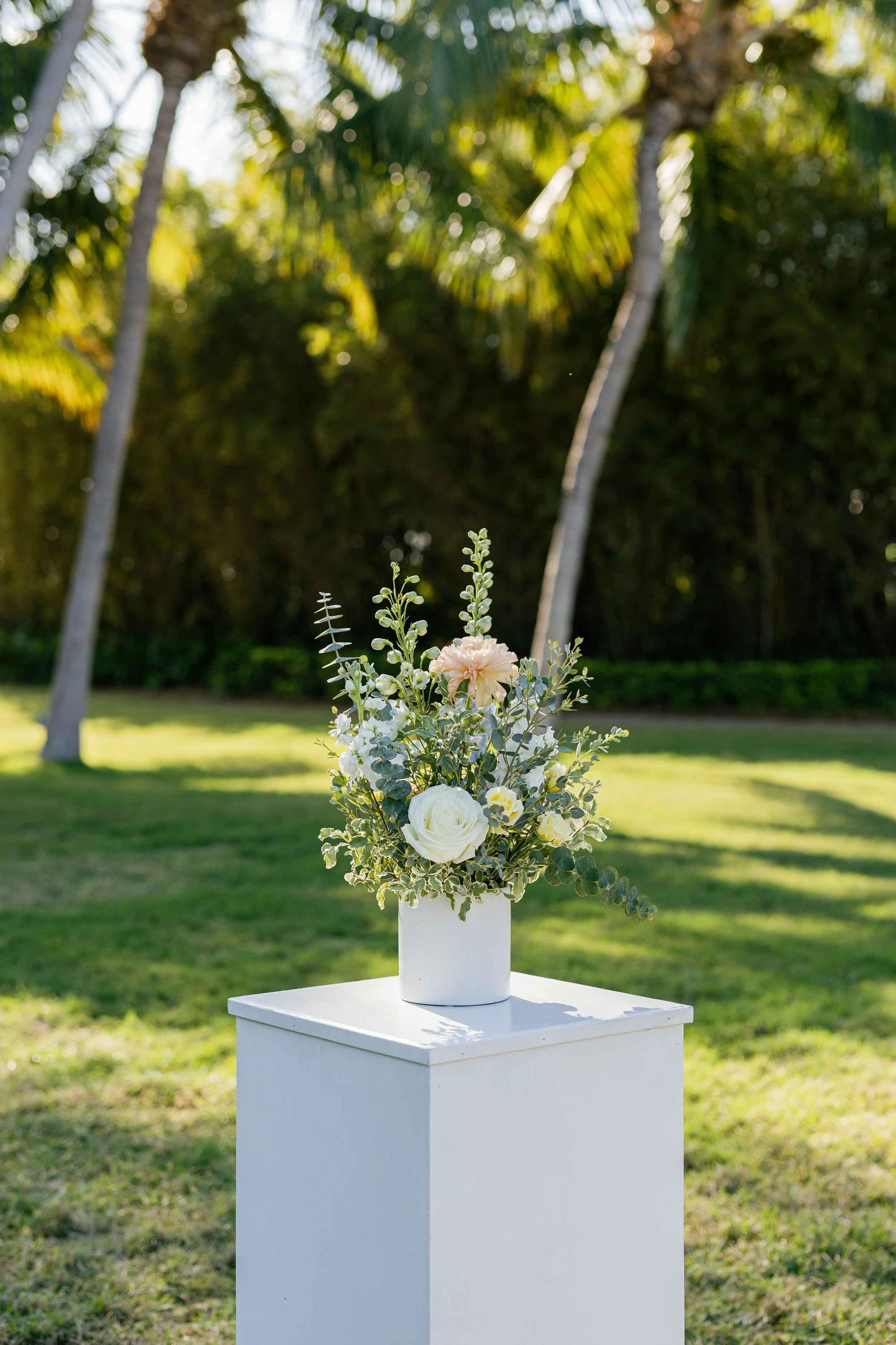 White floral arrangement in a vase placed on a white pedestal outdoors with tall trees and green grass in the background.