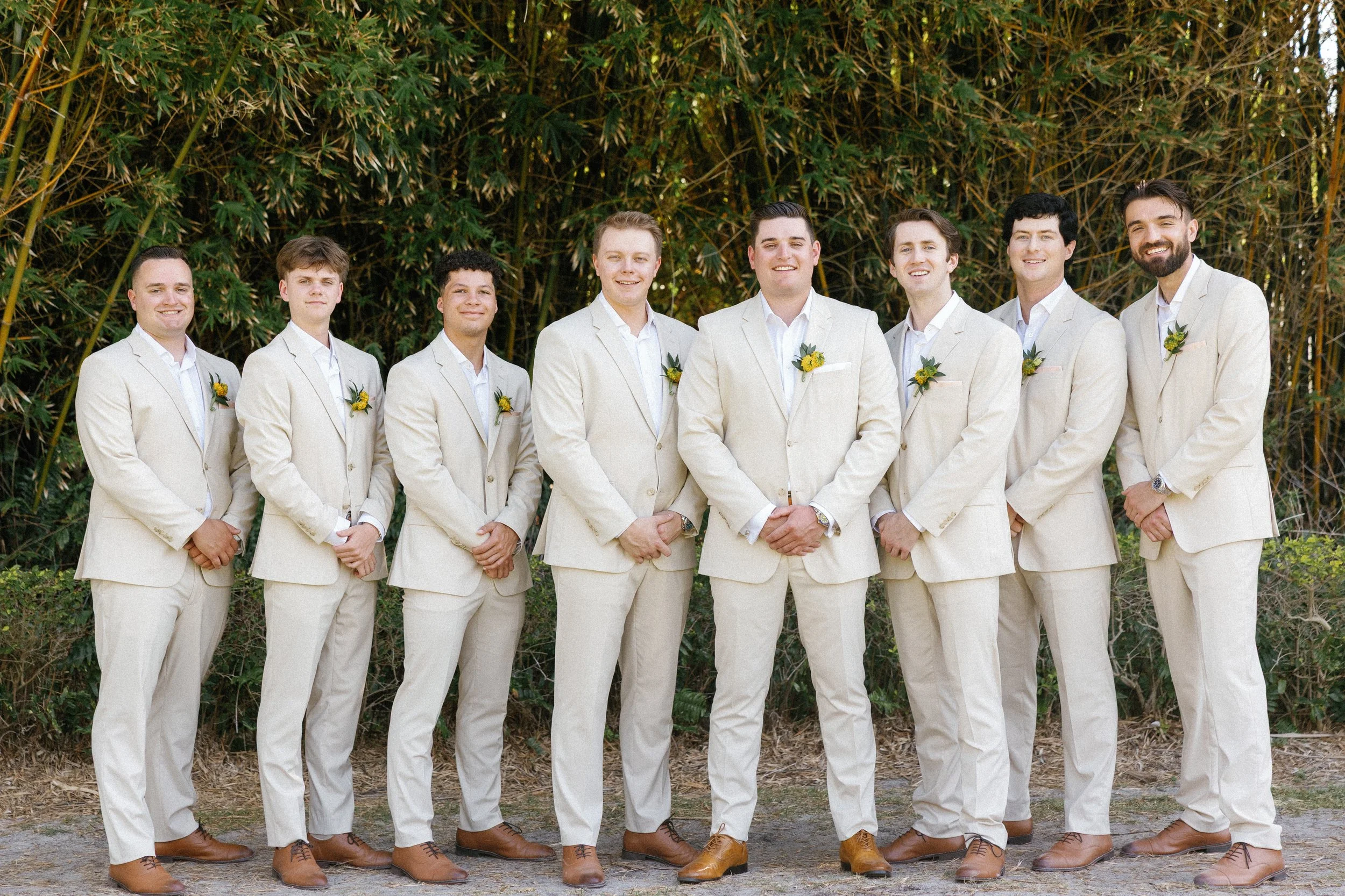A group of nine men dressed in beige suits and white shirts standing outdoors in front of tall green plants, smiling for a photo.