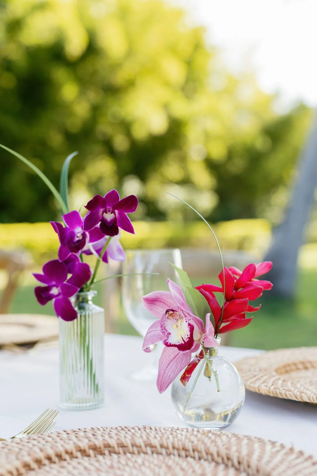 Vases with purple and pink orchids on a table outdoors with a blurred green background.