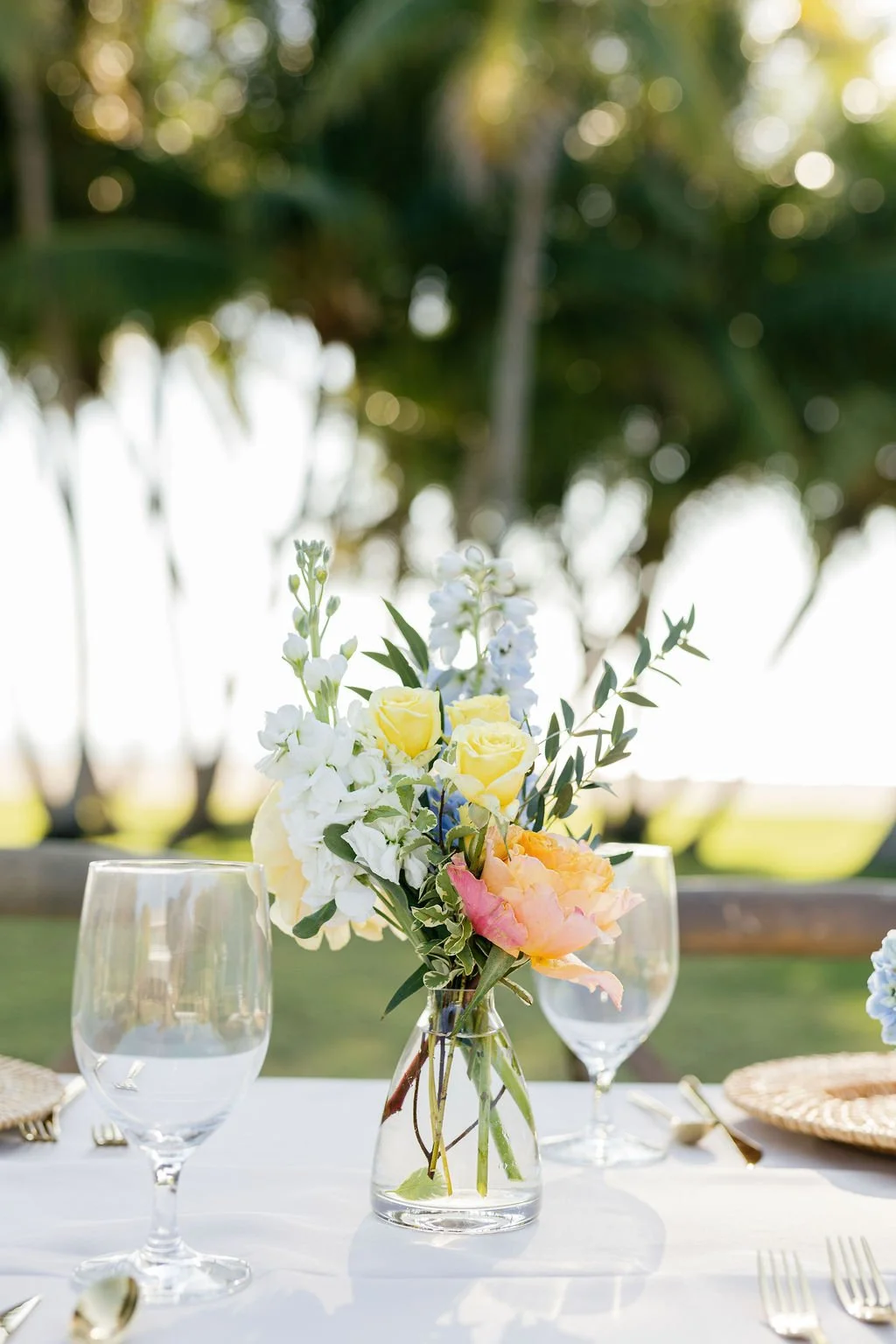 A centerpiece with yellow roses, white flowers, and greenery in a clear glass vase on a white tablecloth, outdoor setting with trees and sunlight in background.