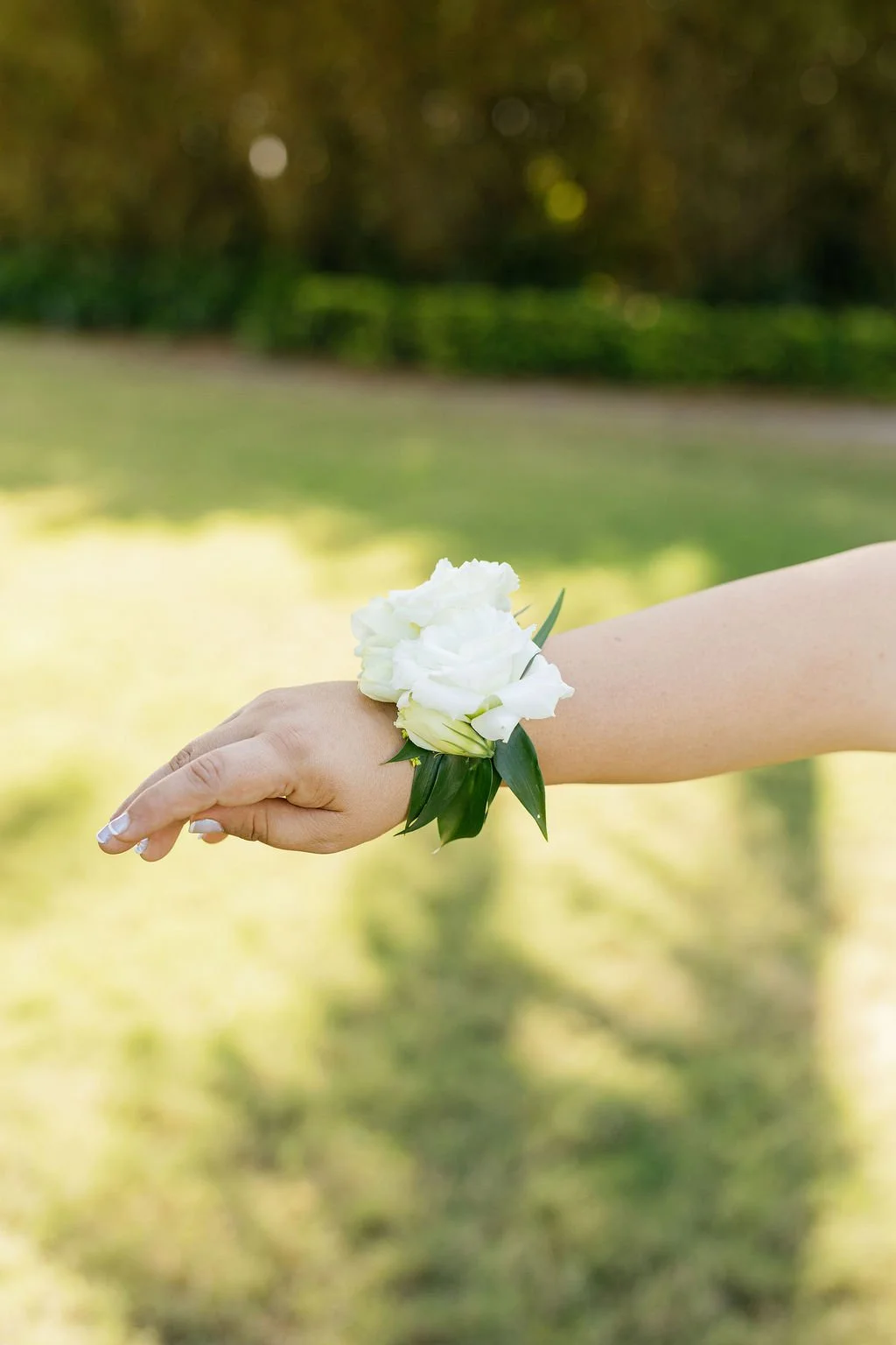 A person's hand wearing a white corsage with white flowers and green leaves, outdoors with a blurred green background.