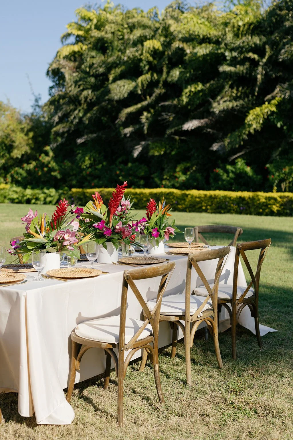 Outdoor banquet table set with glassware, woven placemats, and colorful floral centerpieces, surrounded by wooden chairs on a grassy lawn with trees in the background.