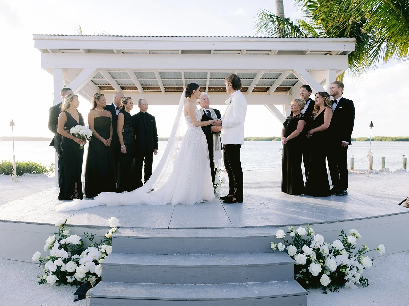 A wedding ceremony taking place outdoors by the water, with a bride in a white dress and veil standing with the groom in a white suit. Bridesmaids and groomsmen dressed in black attire are present, and white flowers decorate the stage area.