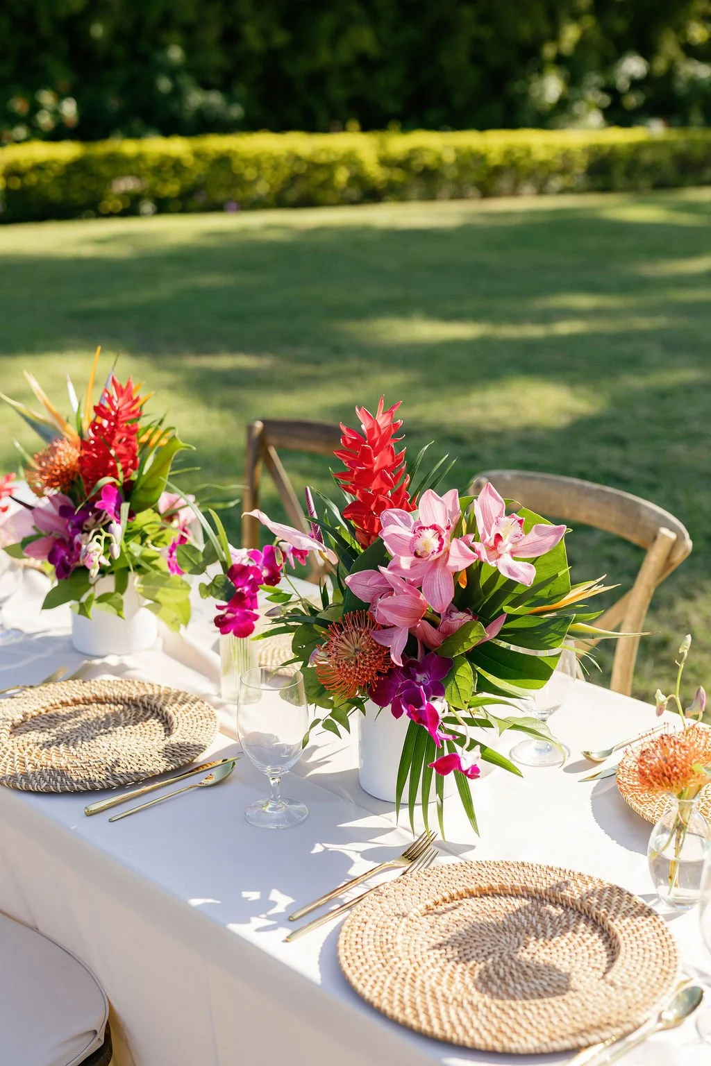Outdoor dining table with colorful floral centerpieces on a white tablecloth, set with woven placemats, glassware, and cutlery, in a sunny garden setting.