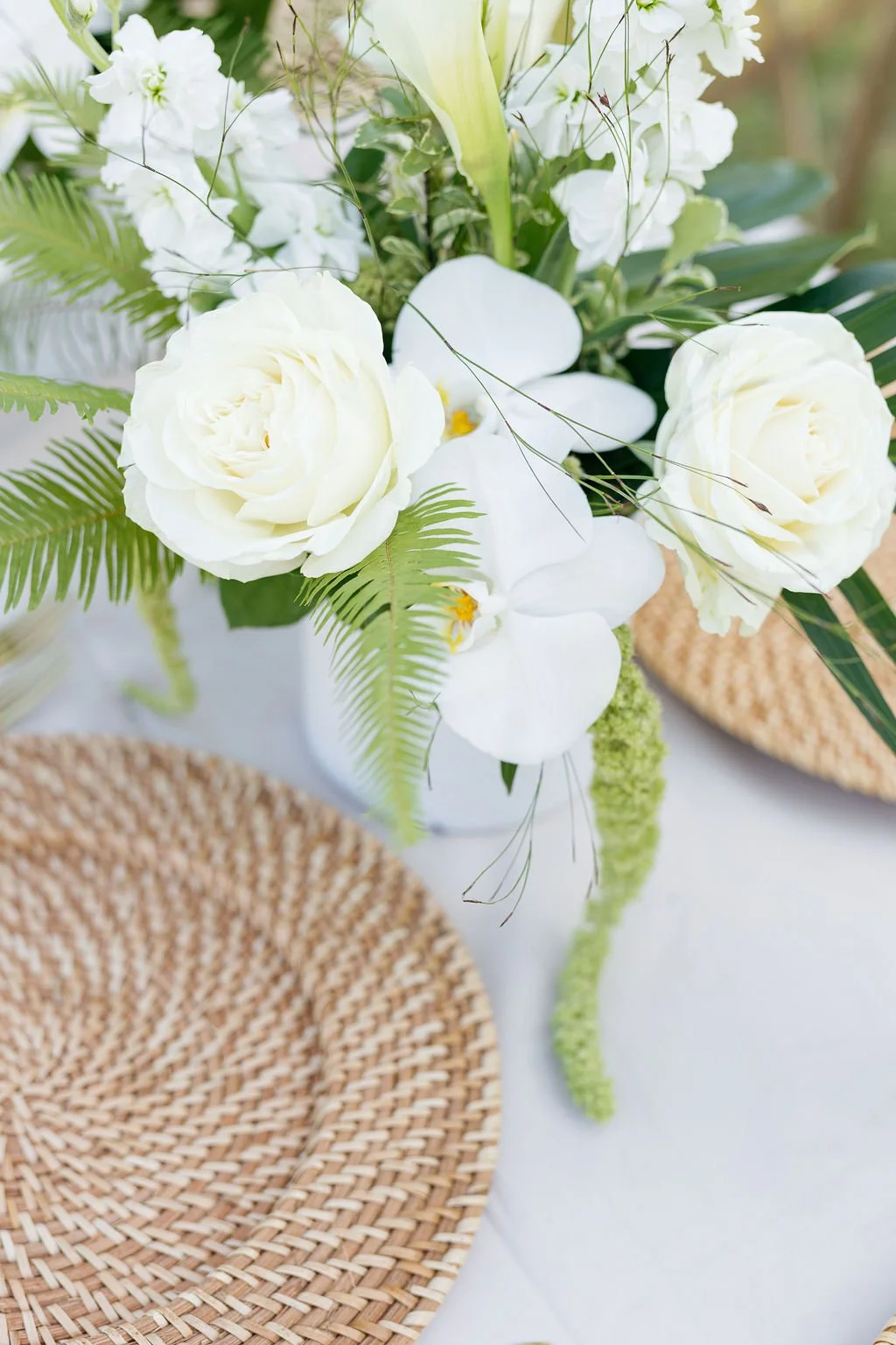 A floral arrangement with white roses, white orchids, green ferns, and other greenery in a white vase, placed on a white table next to a woven rattan placemat.