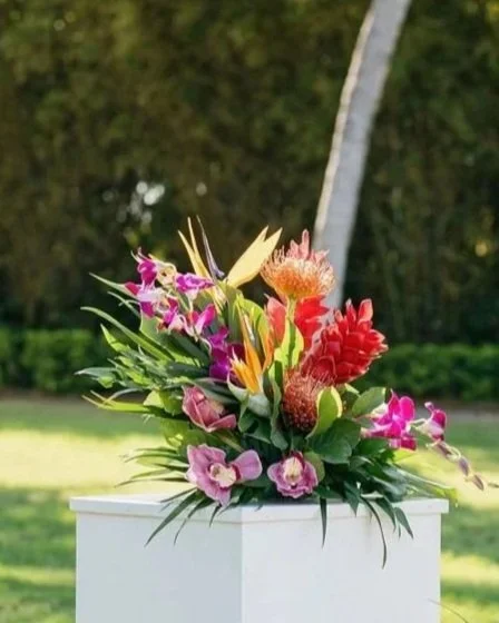 Colorful tropical flower arrangement in a white vase outdoors with greenery and trees in the background.