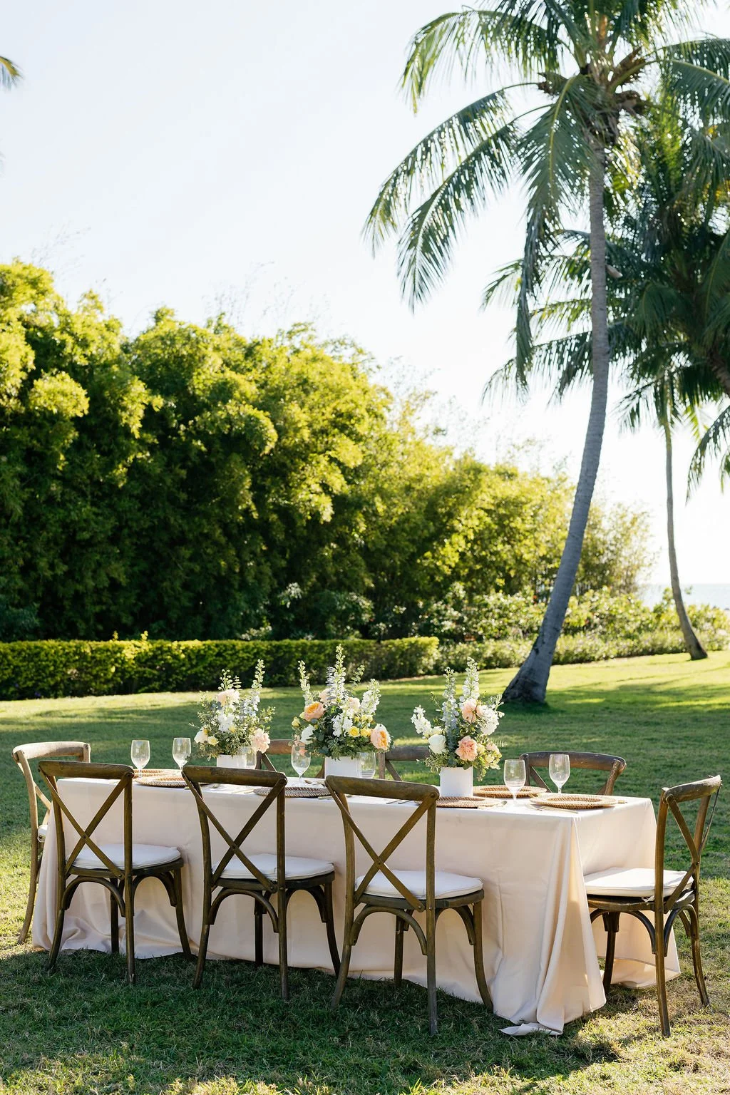 Outdoor table setting with white tablecloth, floral centerpieces, and glassware, under palm trees in a lush green garden.