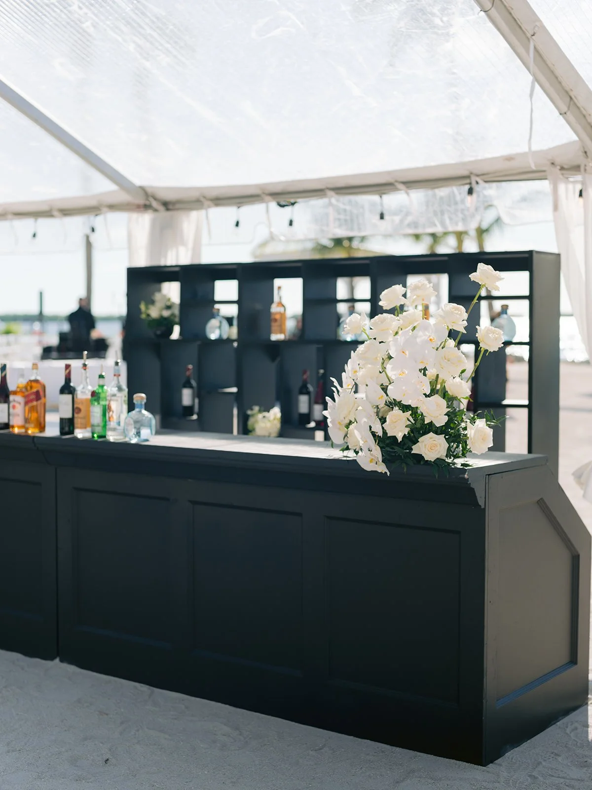 Outdoor bar with black counter, bottles of liquor, and white flower arrangement under a transparent tent canopy.