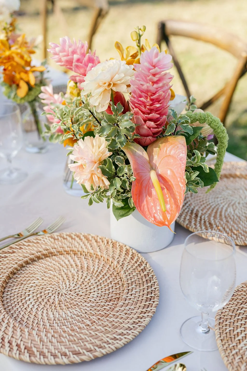 A floral centerpiece with pink and white flowers, including anthuriums, set on a dining table with woven placemats and glassware.