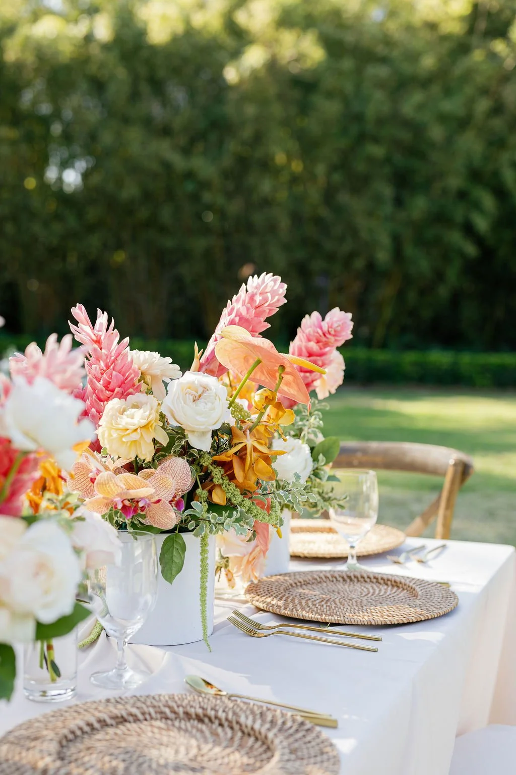 Elegant outdoor table setting with a large floral centerpiece in a white vase, featuring pink, white, and orange flowers, with woven placemats and gold utensils, on a white tablecloth, set in a lush green garden.