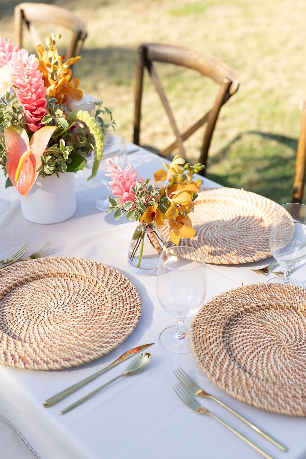 Elegant outdoor dining table with woven chargers, gold utensils, clear wine glasses, and floral centerpieces in pink, yellow, and green colors on a white tablecloth.