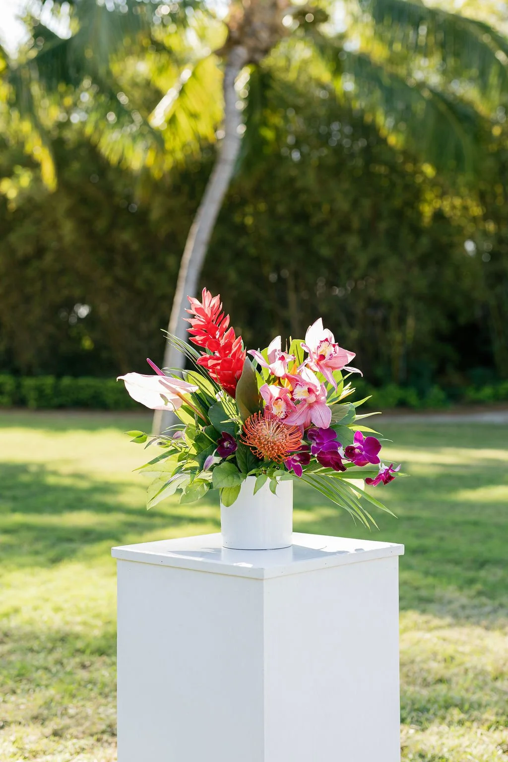 A colorful flower arrangement in a white vase on a white pedestal outdoors, with a background of green trees and grass.