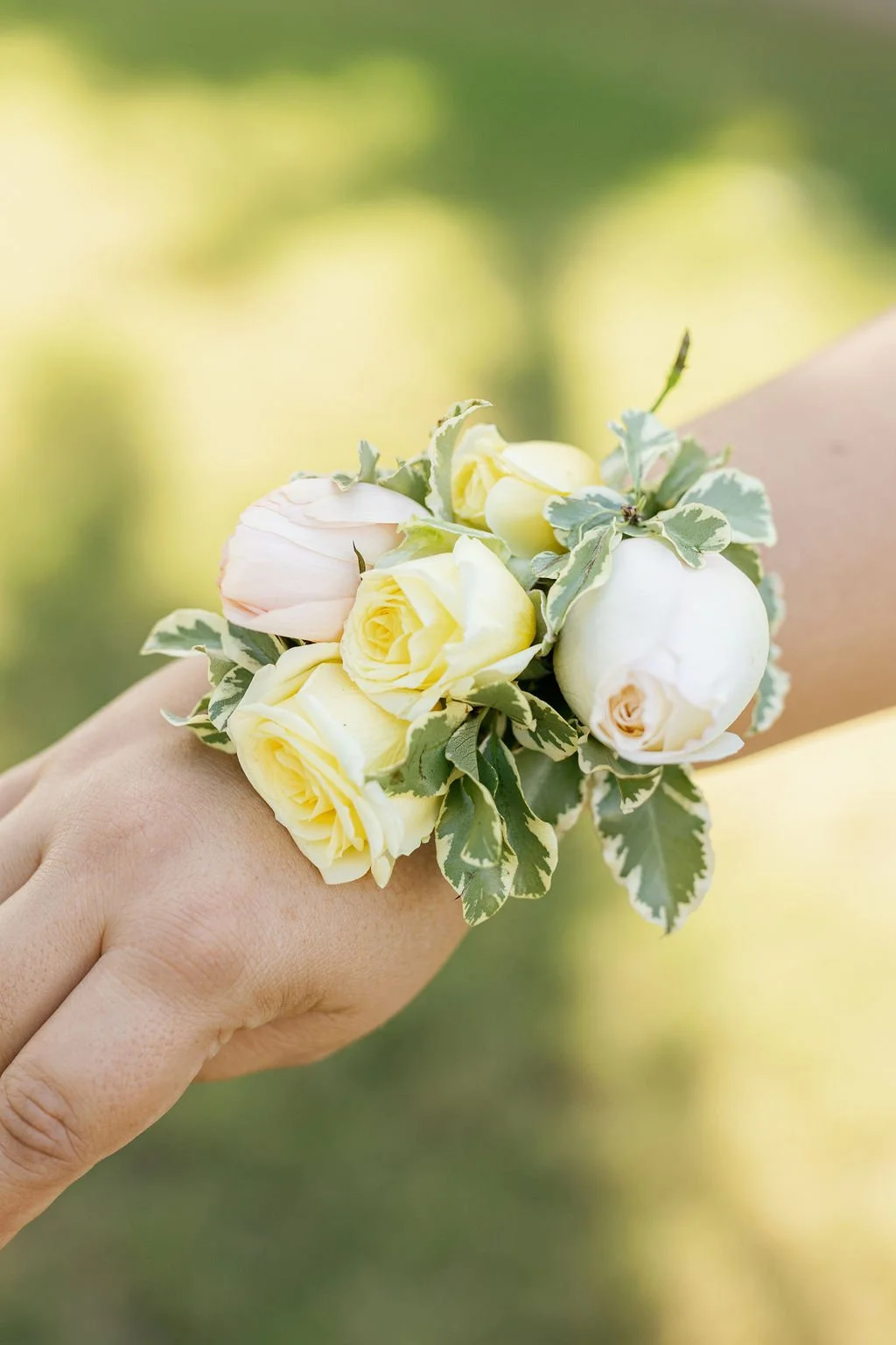 A person's hand holding a small bouquet of yellow roses, white peonies, and variegated green and white leaves.