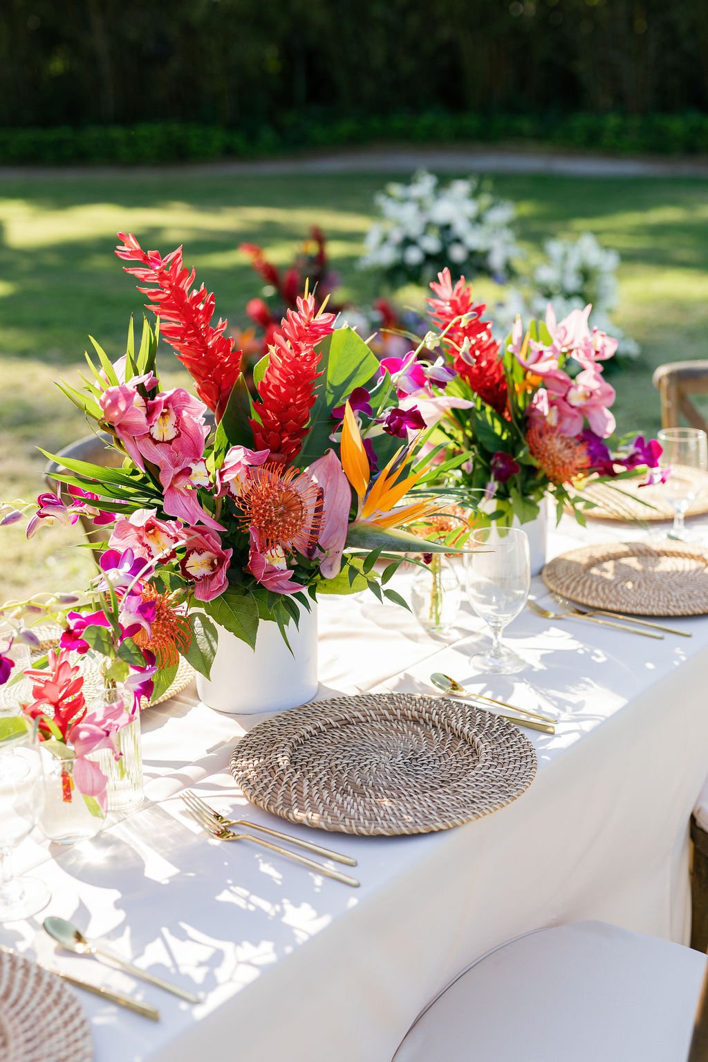 A table set for outdoor dining with colorful floral centerpieces, glassware, and place settings on a white tablecloth, with a grassy area and trees in the background.