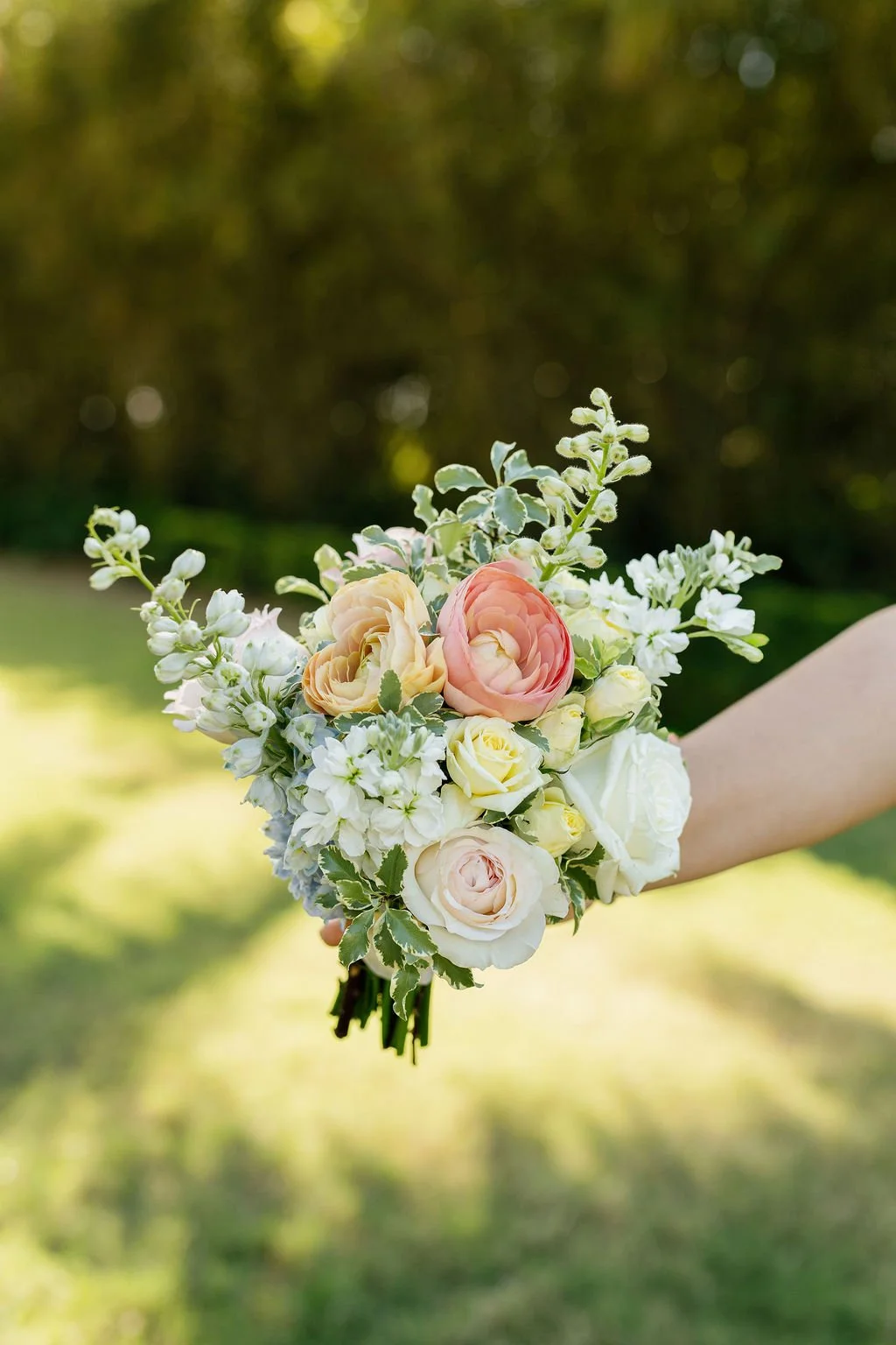 A hand holding a bouquet of mixed flowers, including roses and ranunculus, with a blurred outdoor background.