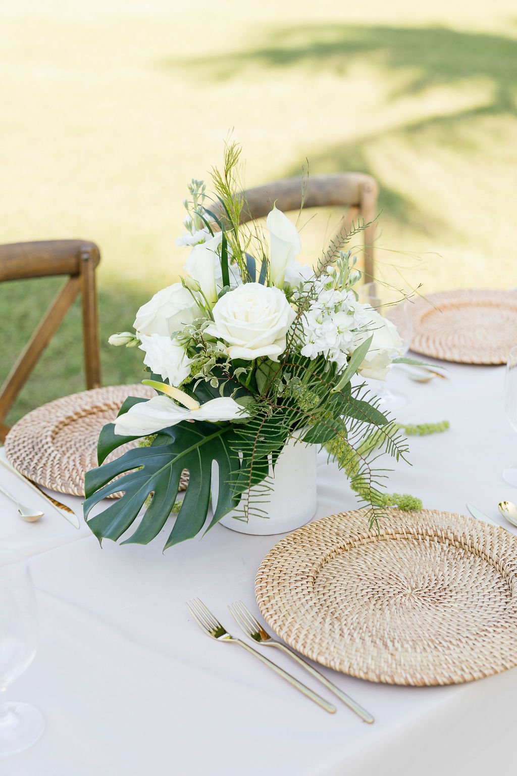 A table set for outdoor dining with a centerpiece of white flowers including roses, calla lilies, and greenery in a white vase, surrounded by woven placemats, gold flatware, and white tablecloths, with a grassy background.