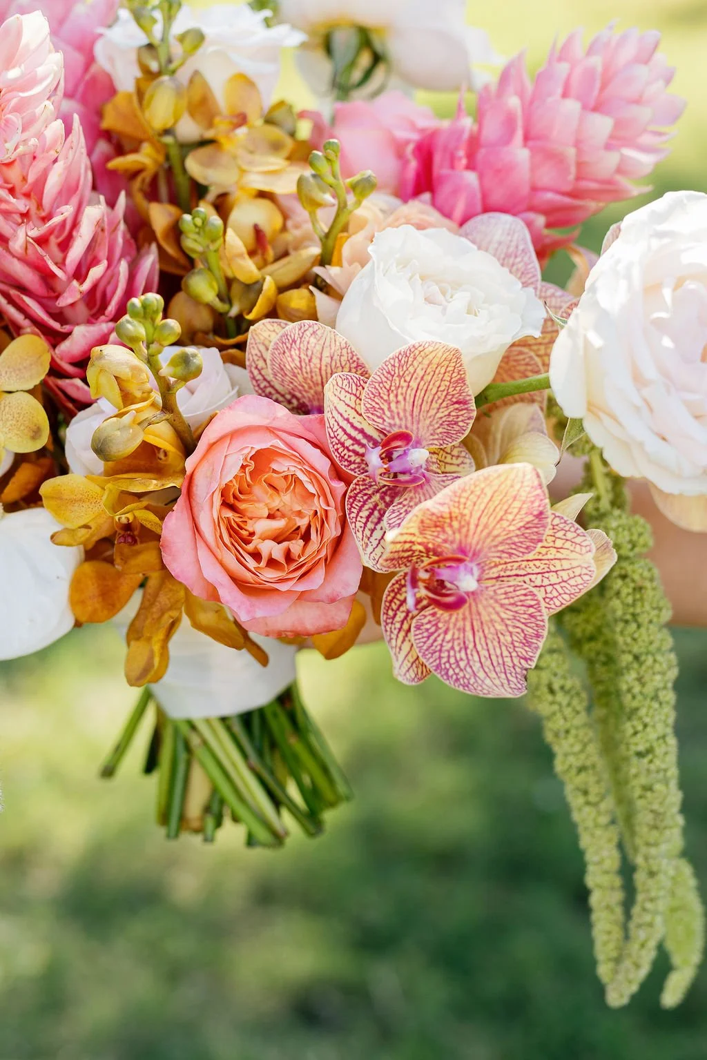 Close-up of a colorful flower bouquet featuring pink roses, white roses, yellow orchids, pink ginger, and striped orchids against a blurred green background.