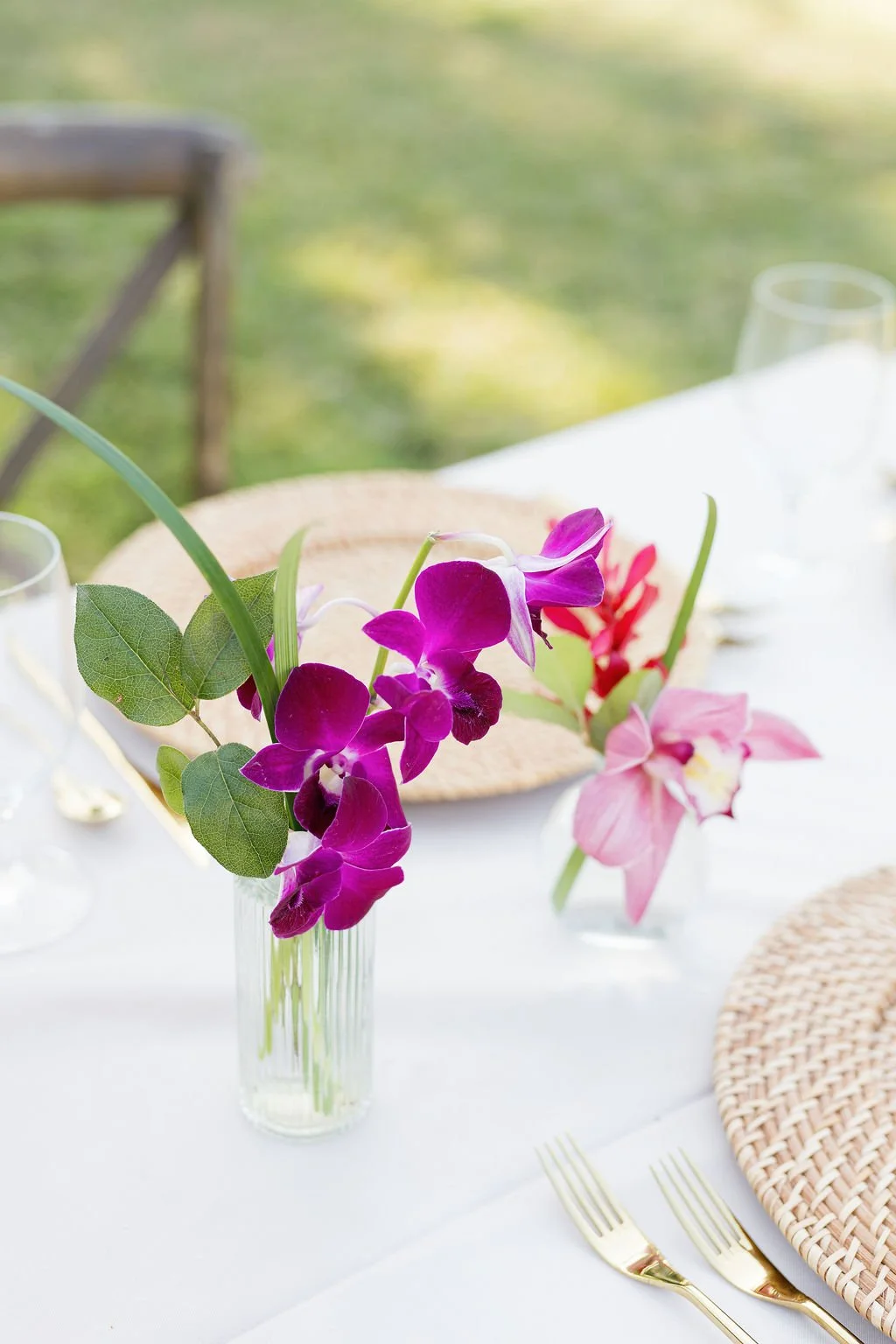 A table decorated with a vase of purple and pink flowers, set outdoors with woven placemats, gold utensils, and glassware.