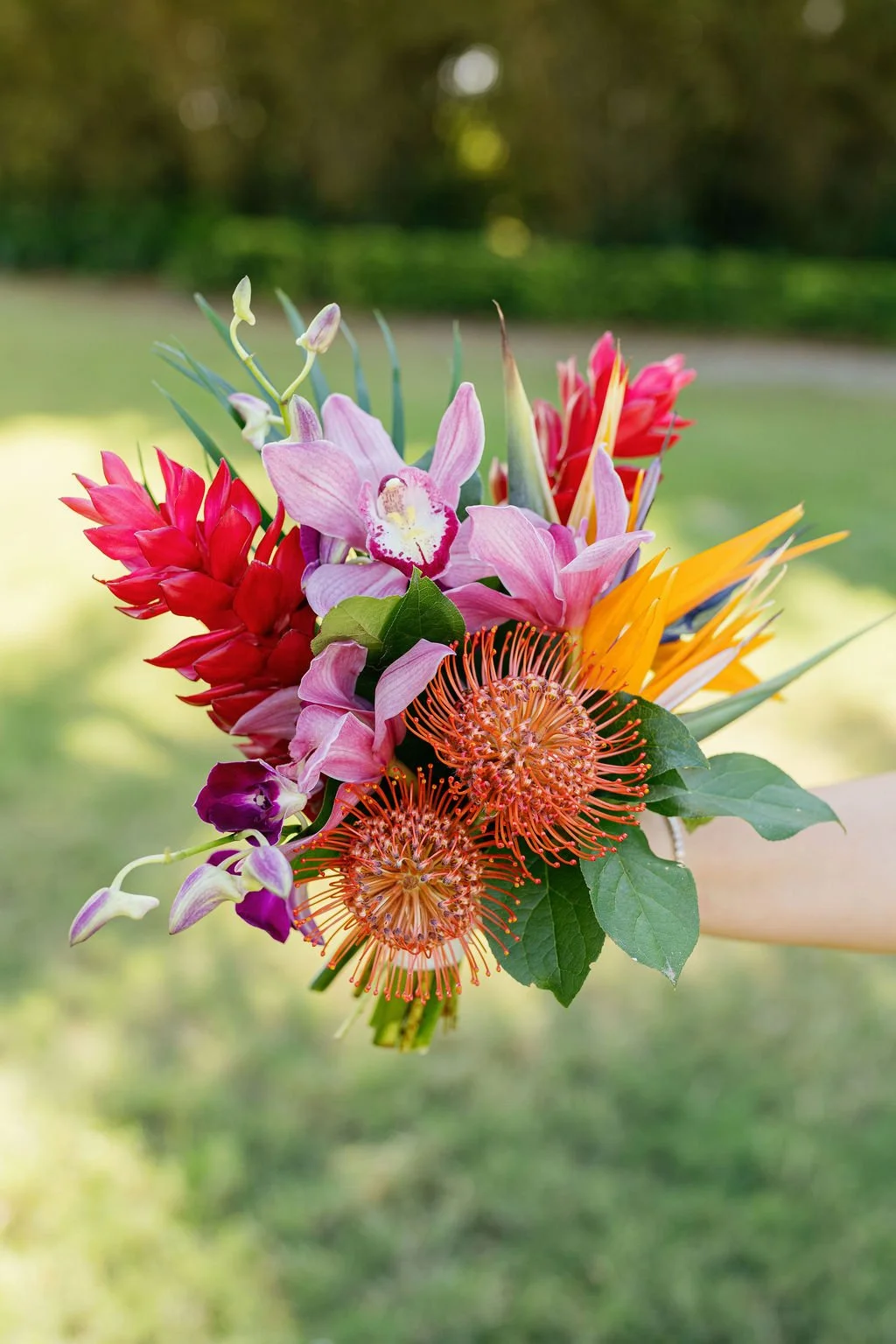 Colorful bouquet of various flowers, including pink orchids, red ginger, orange pincushion protea, and yellow bird of paradise, held outdoors with blurred green background.