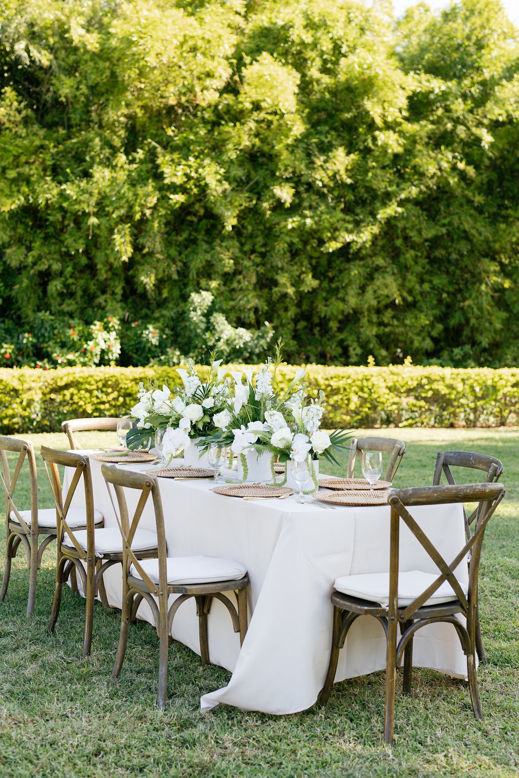 Outdoor dining table decorated with white floral centerpiece, set with placemats, wine glasses, and white chairs, placed on grass with trees and a hedge in background.