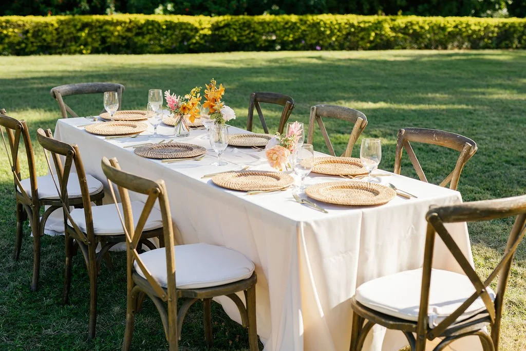 Outdoor dining table set for eight, with a white tablecloth, wicker plates, wine glasses, and floral centerpieces, on a grassy lawn.