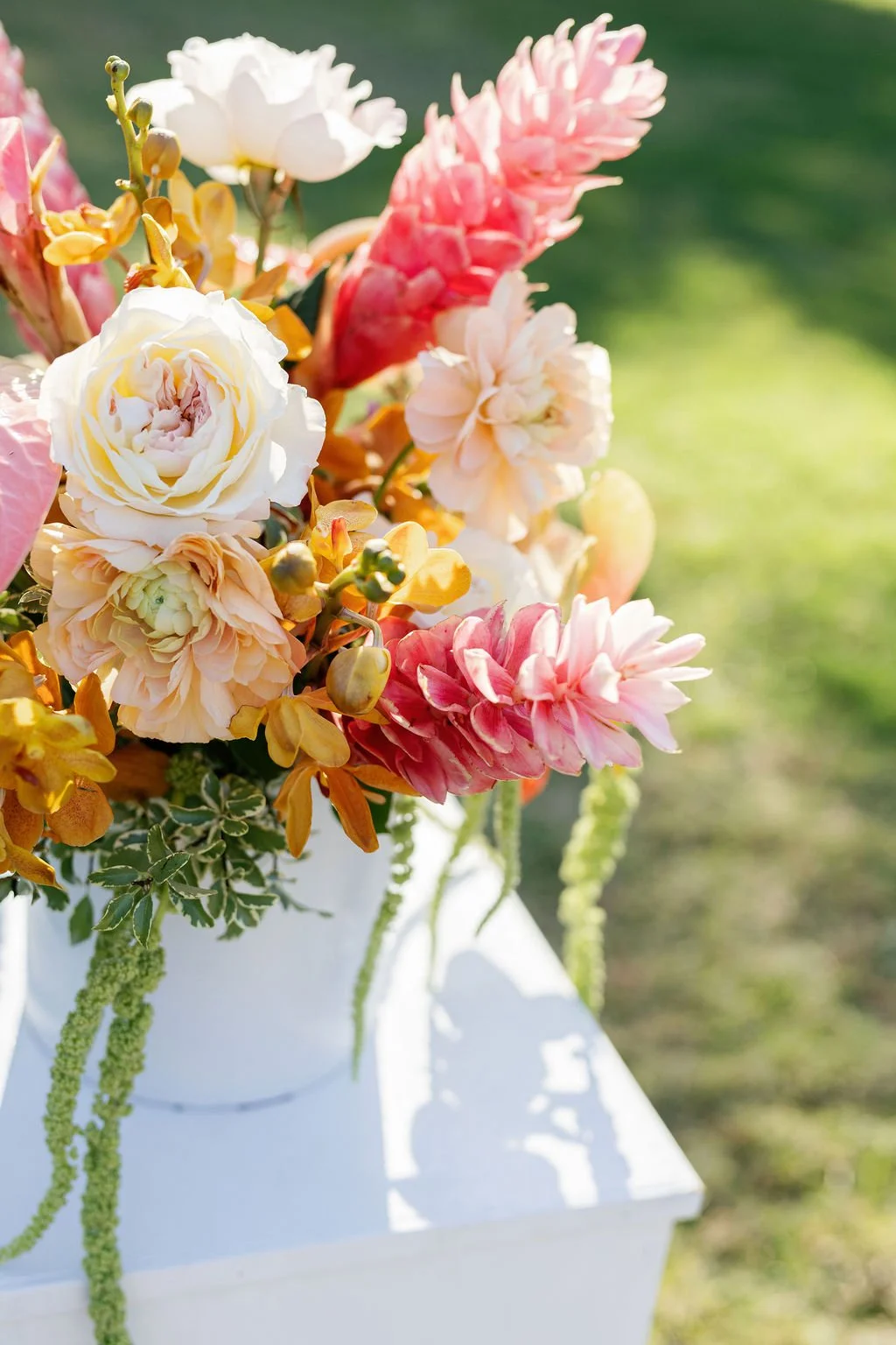 A colorful bouquet of pink, white, peach, and yellow flowers in a white vase outdoors, with a blurred green background.