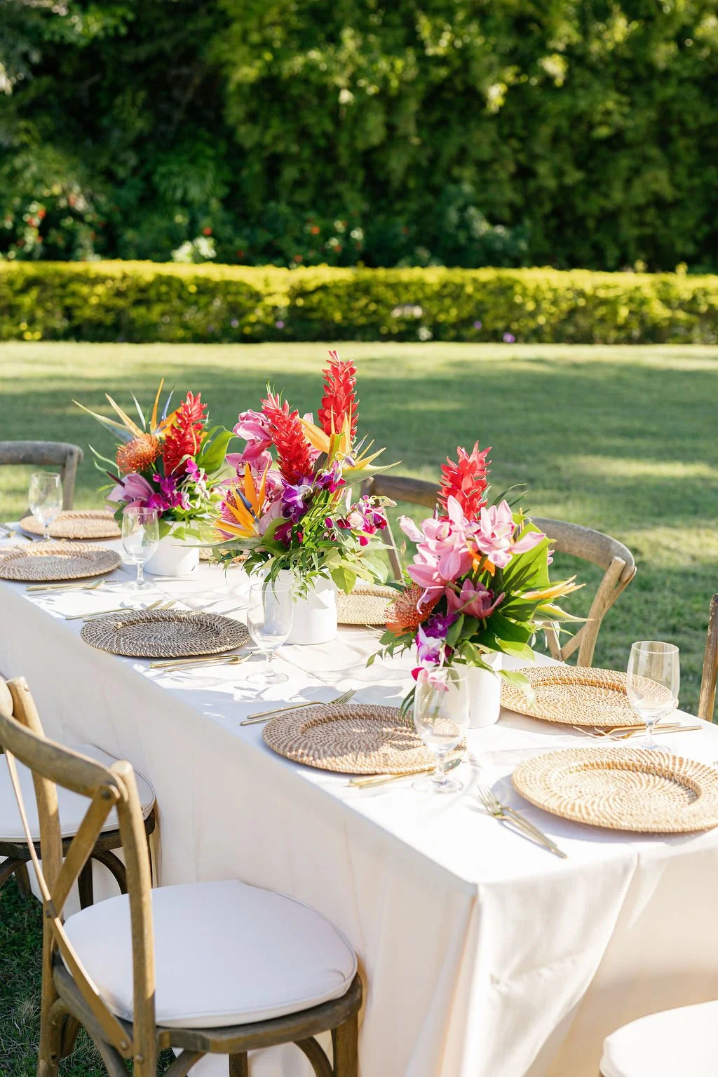 Outdoor table setup with colorful floral arrangements, glassware, and woven placemats in a lush green garden.