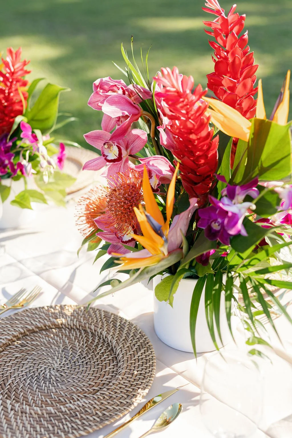 A vibrant flower arrangement with pink orchids, red ginger, orange protea, and purple orchids in a white vase on a table outdoors.