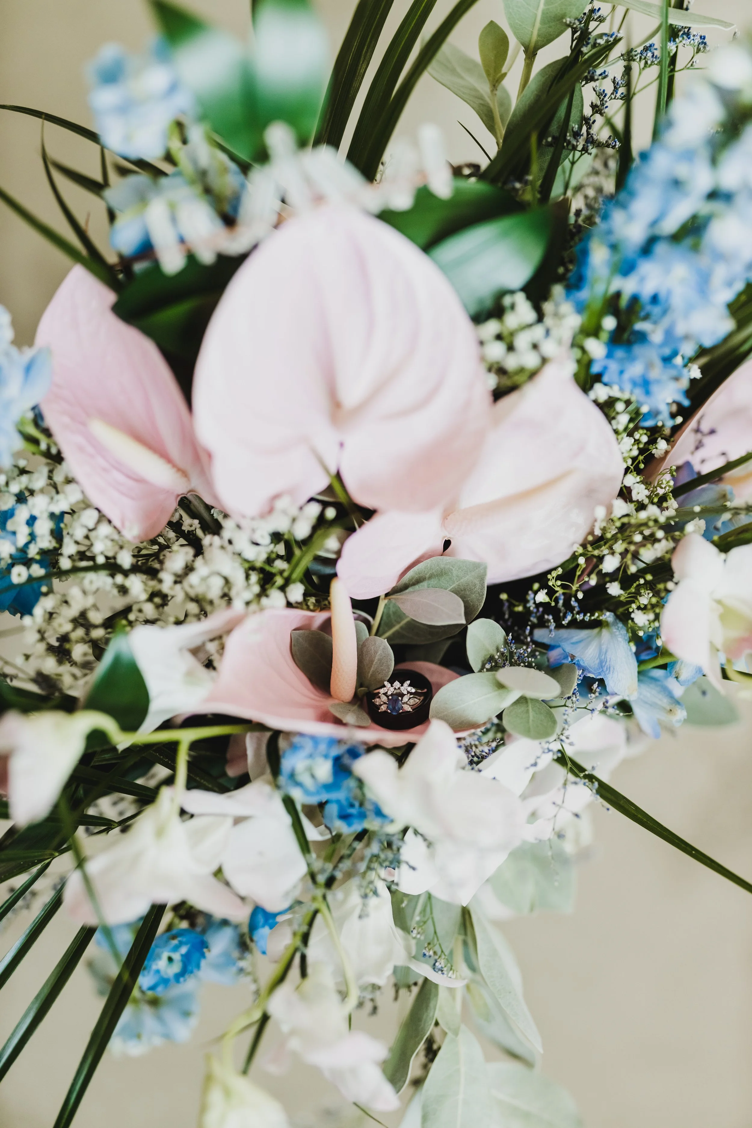 Bouquet with pink anthuriums, blue delphiniums, greenery, and a diamond ring.