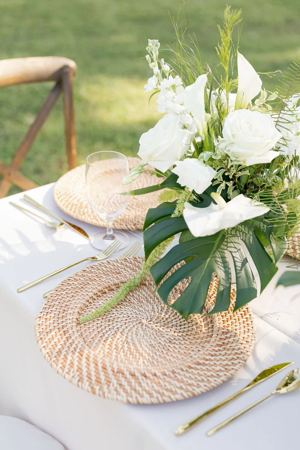 Elegant outdoor table setting with a bouquet of white roses and greenery on woven placemats, gold utensils, and a wine glass