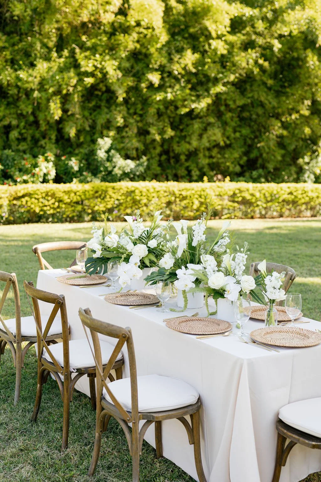 Outdoor table set for a celebration, decorated with white floral arrangements, glassware, and woven placemats, surrounded by wooden chairs on a grassy lawn.