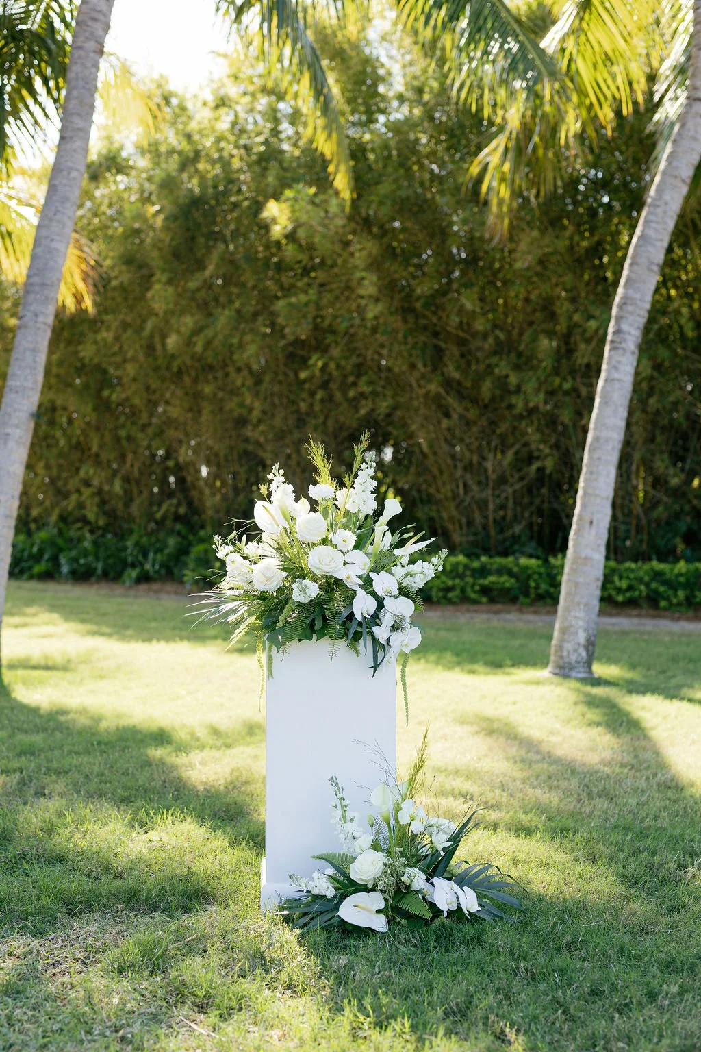 White floral arrangement on a white pedestal outdoors, with palm trees and greenery in the background.