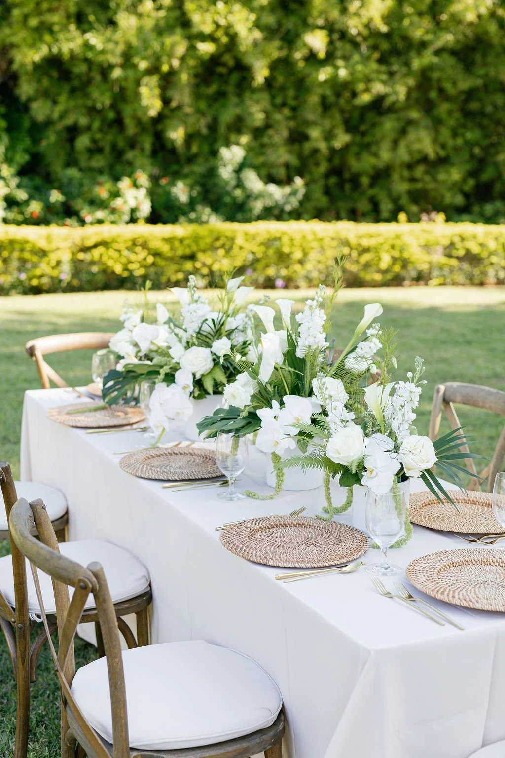 Outdoor table decorated with white floral centerpieces, wicker placemats, glassware, and gold-toned cutlery, set in a garden with green hedges and trees in the background.