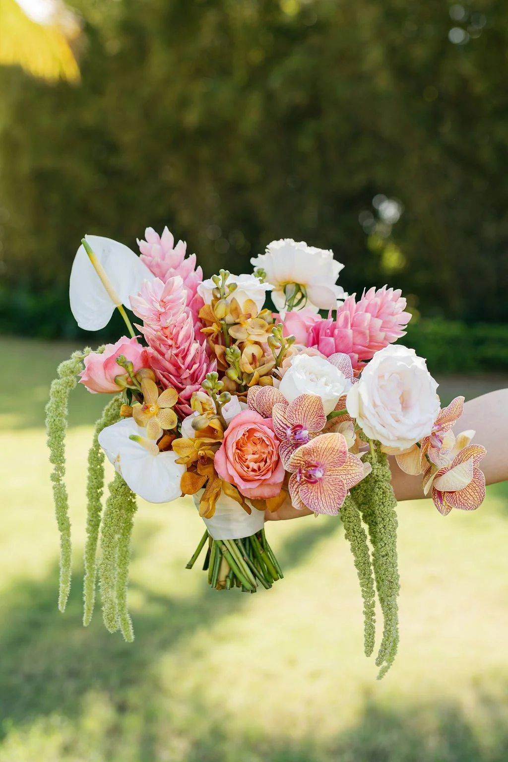 A hand holding a bouquet of various pink, white, and peach-colored flowers with hanging green accents, set against a blurred outdoor background.