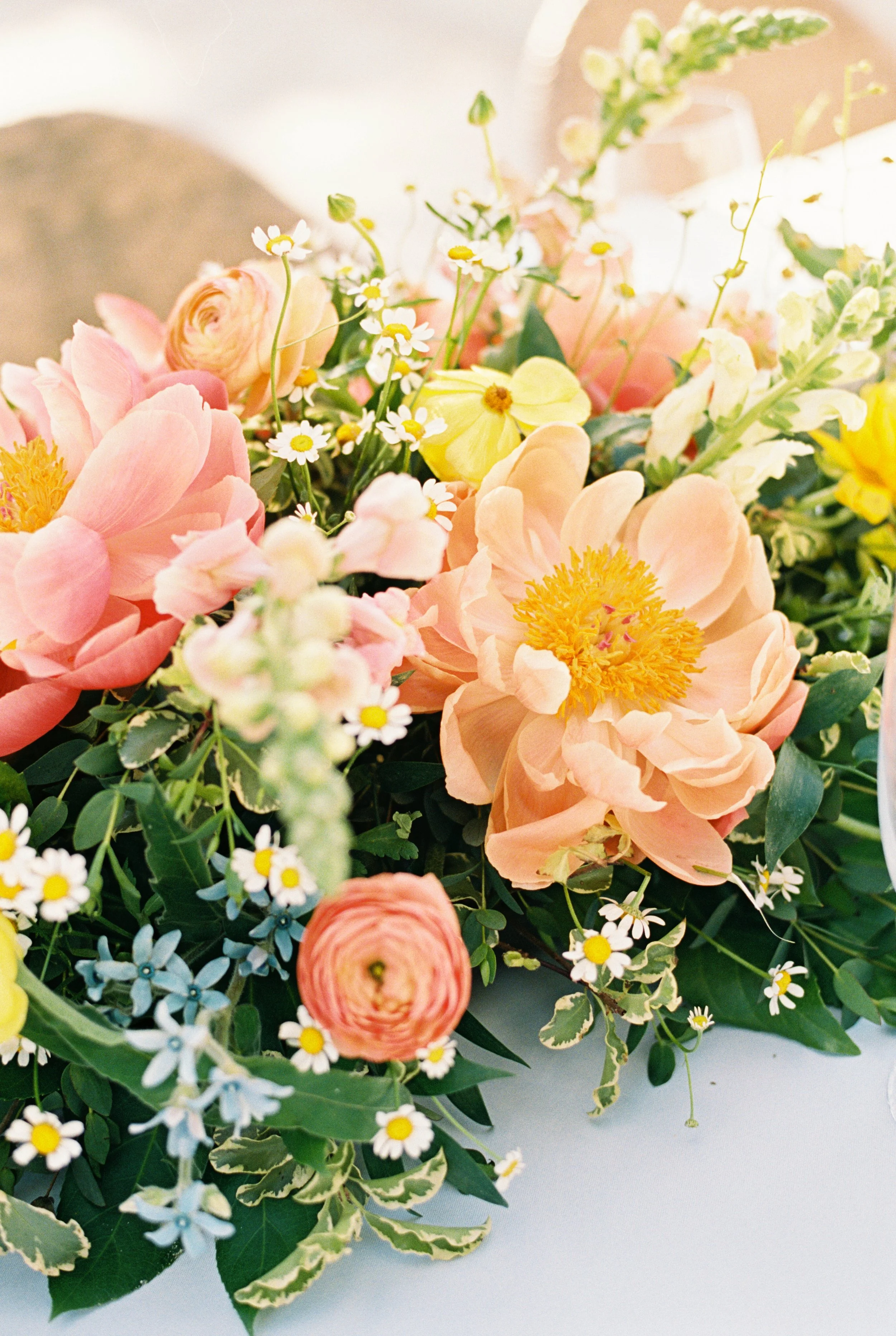 Close-up of a colorful flower arrangement with pink peonies, yellow poppies, small white daisies, and blue forget-me-nots surrounded by green foliage.