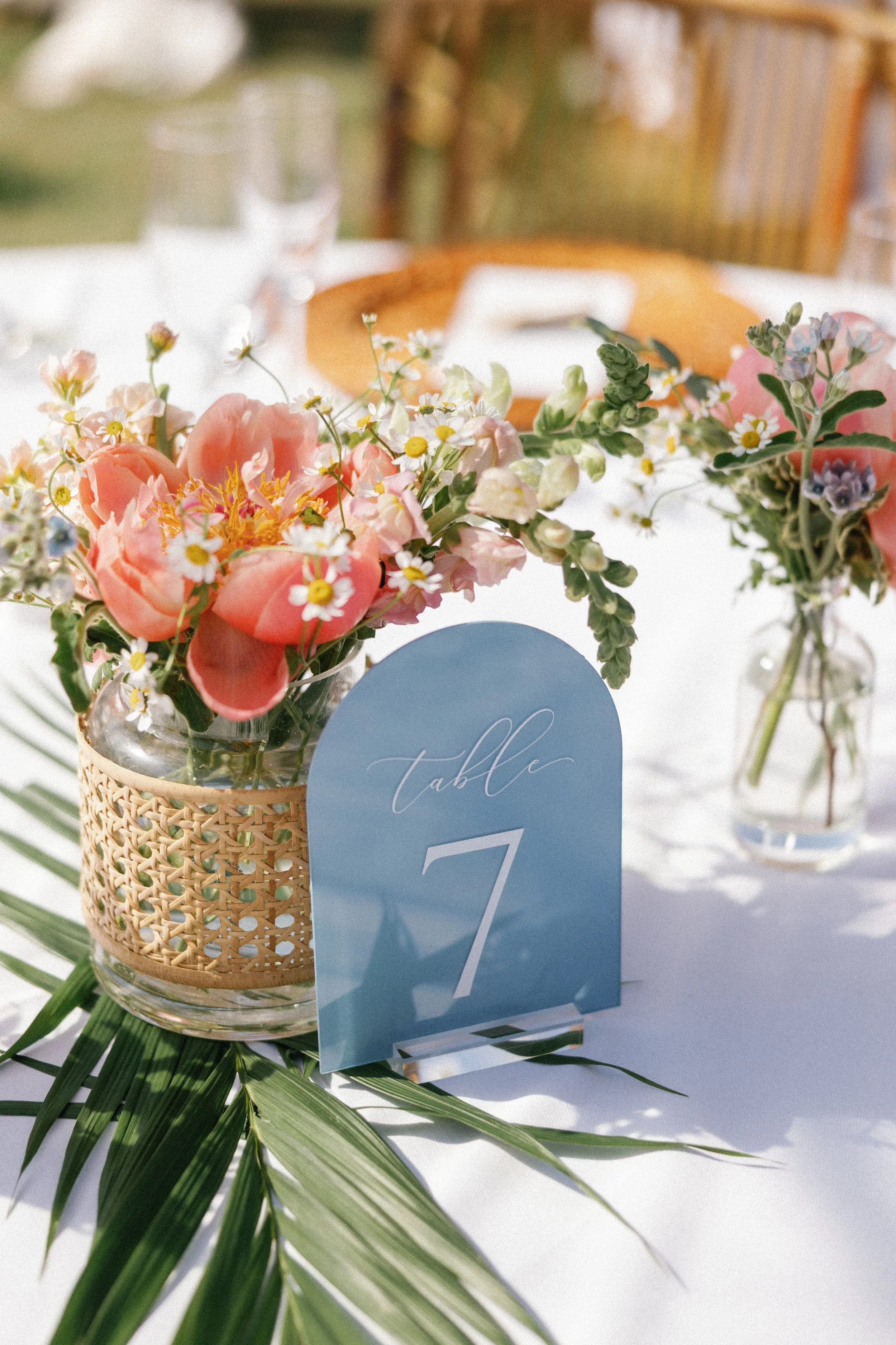 A table centerpiece with pink flowers in a glass vase wrapped in a woven cover, a blue table number card labeled 'table 7', and some greenery beneath, set on a white tablecloth with other glassware in the background.