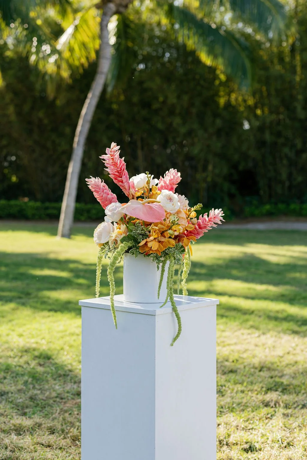 Colorful flower arrangement with pink, white, yellow, and peach flowers in a white vase on a white pedestal outdoors with grassy background and palm trees.